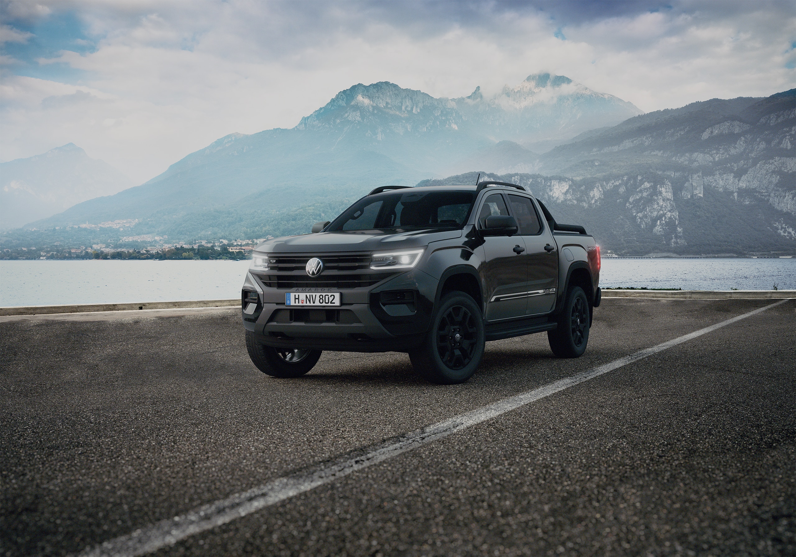Dunkler Volkswagen Amarok steht vor einem See mit Bergen und Häusern im Hintergrund.