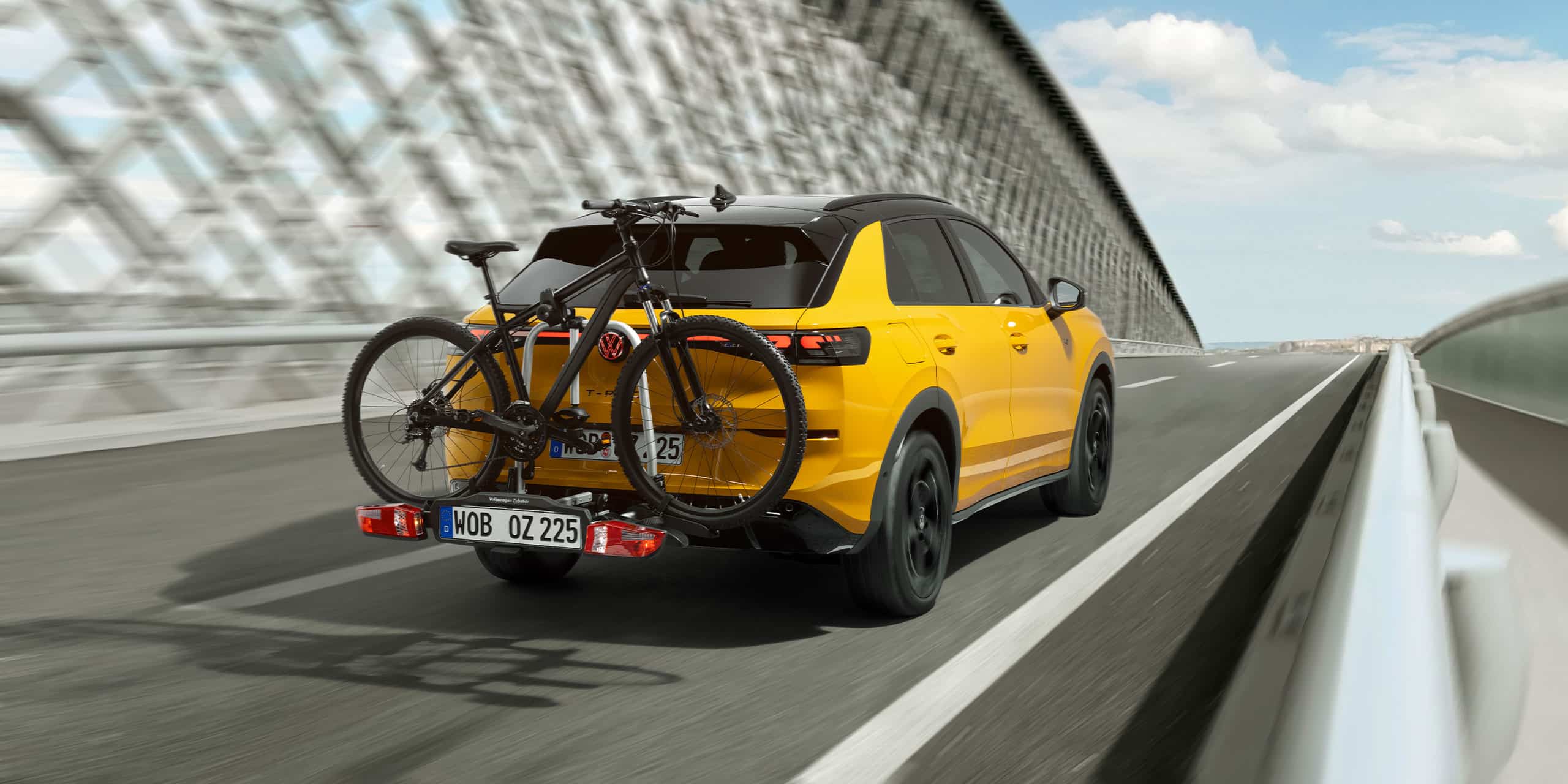 Gelber Volkswagen T-Roc fährt auf einer Brücke und hat hinten am Auto ein Fahrrad montiert.