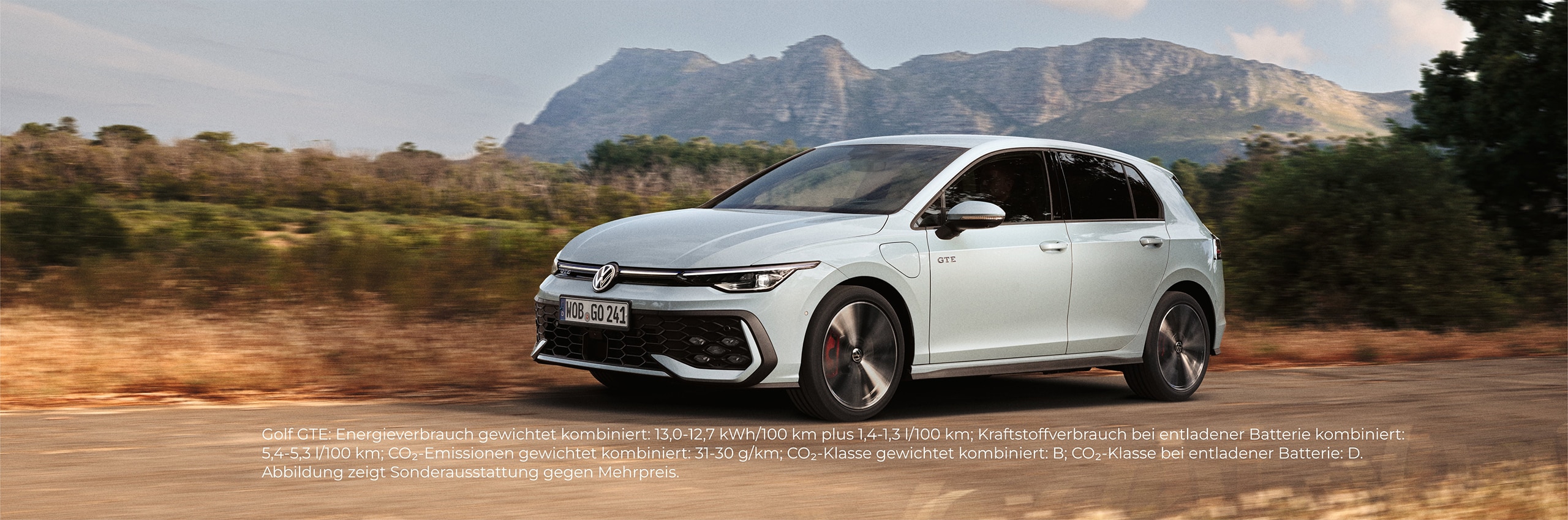 Weißer Volkswagen fährt auf einer Straße mit Bergen im Hintergrund.