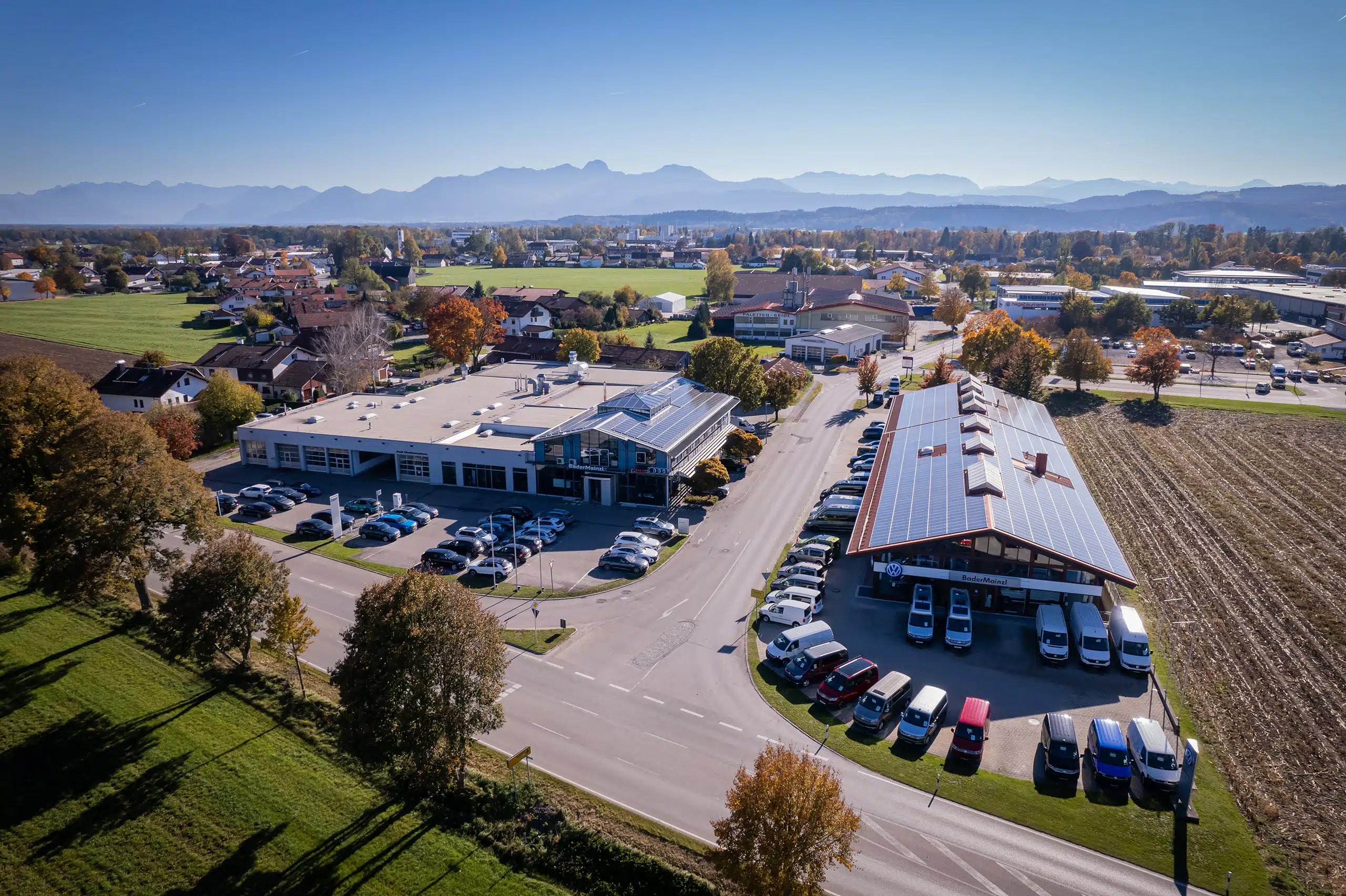 Luftaufnahme des Standorts Autowelt Bader in Bruckmühl-Heufeld: zwei Autohaus-Gebäude mit Solardächern und Parkplätzen, dahinter die Alpen